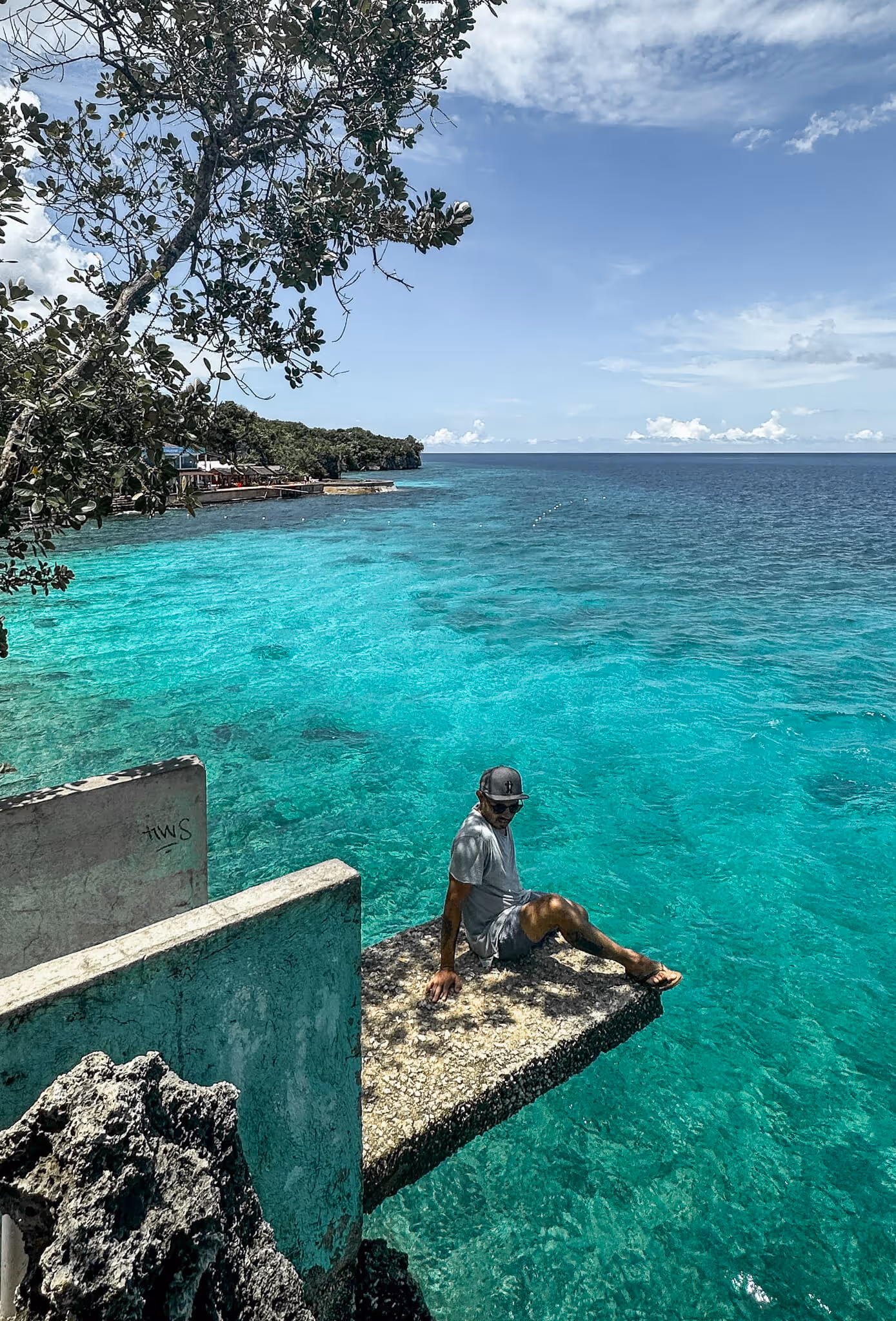 Person sitting on a concrete ledge over clear turquoise ocean water with trees and blue sky.