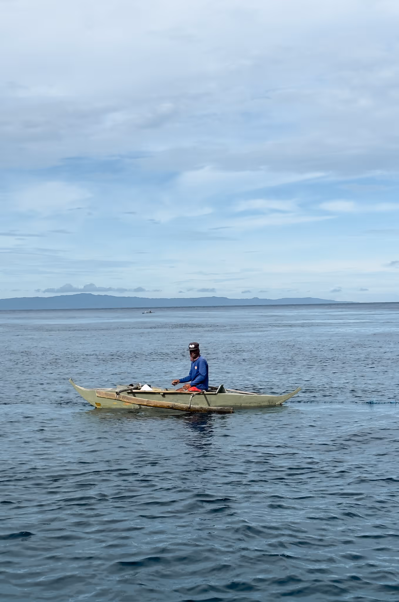 Man in a blue shirt sitting in a small wooden boat on calm ocean water with distant land visible.