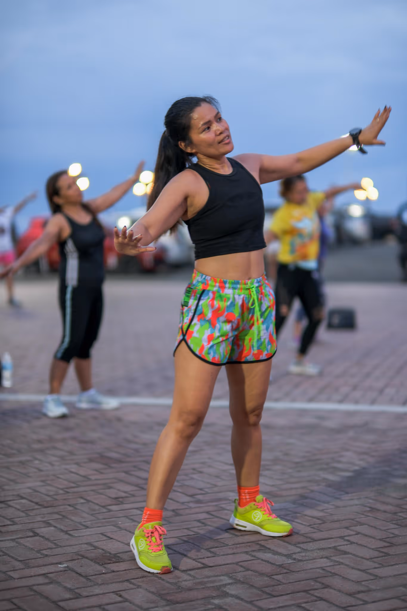 Woman in colorful shorts and black top stretching arms during outdoor group exercise at dusk.