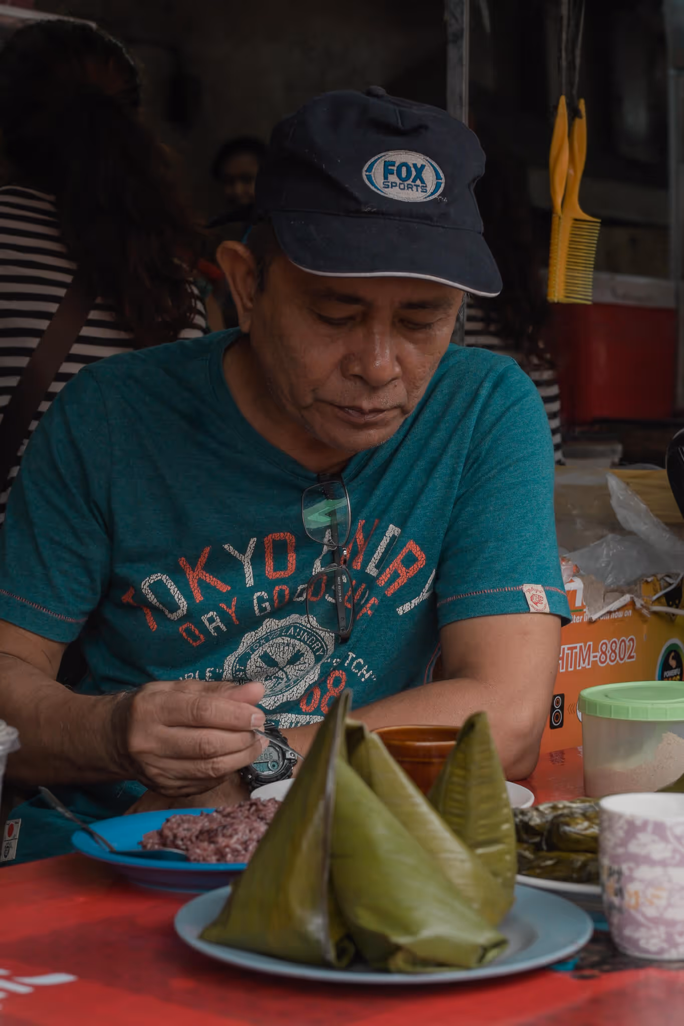 Man in cap eating traditional food wrapped in banana leaves at a red table.