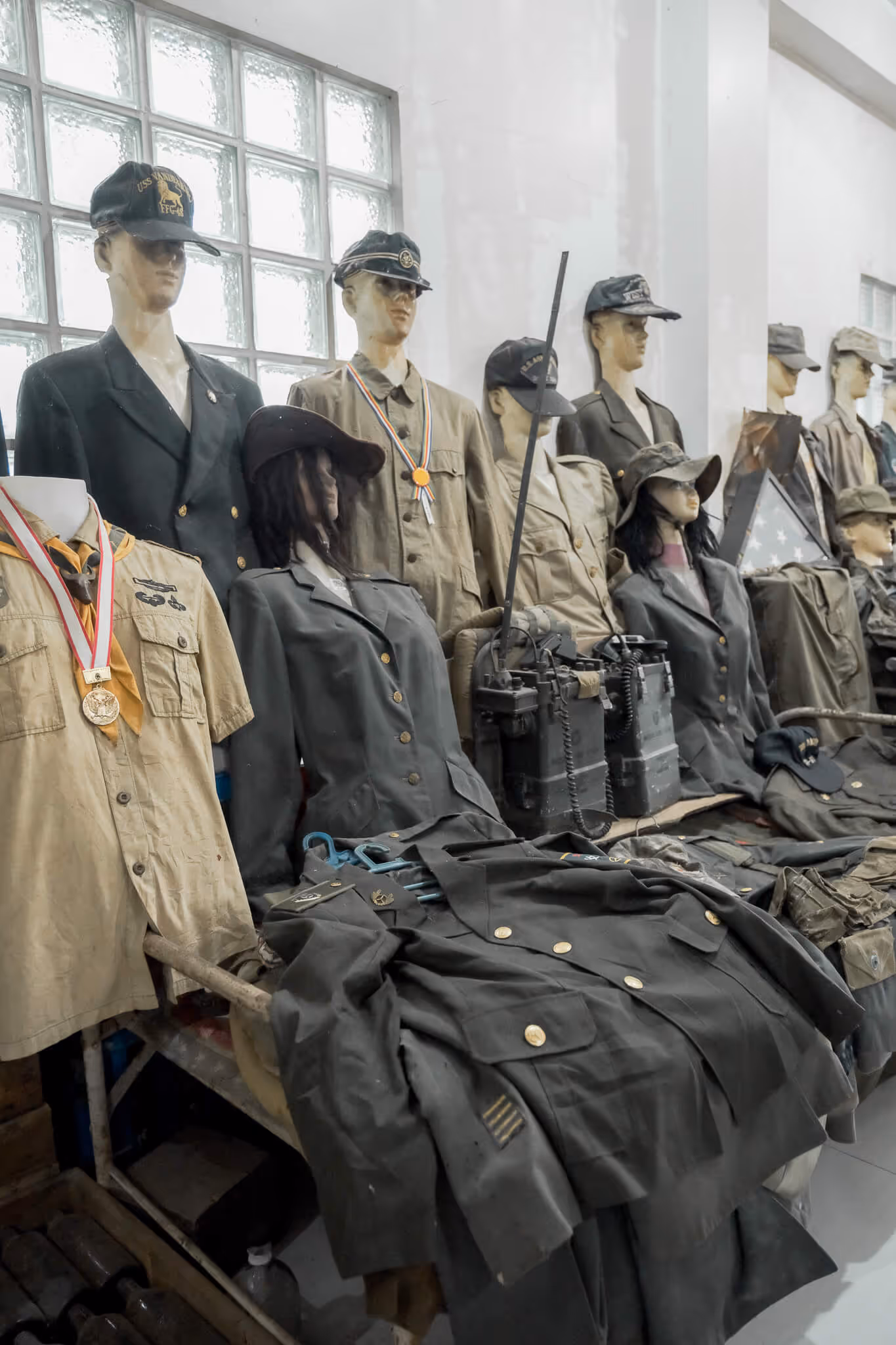 Mannequins wearing vintage military uniforms, hats, and medals displayed indoors near a glass block wall.