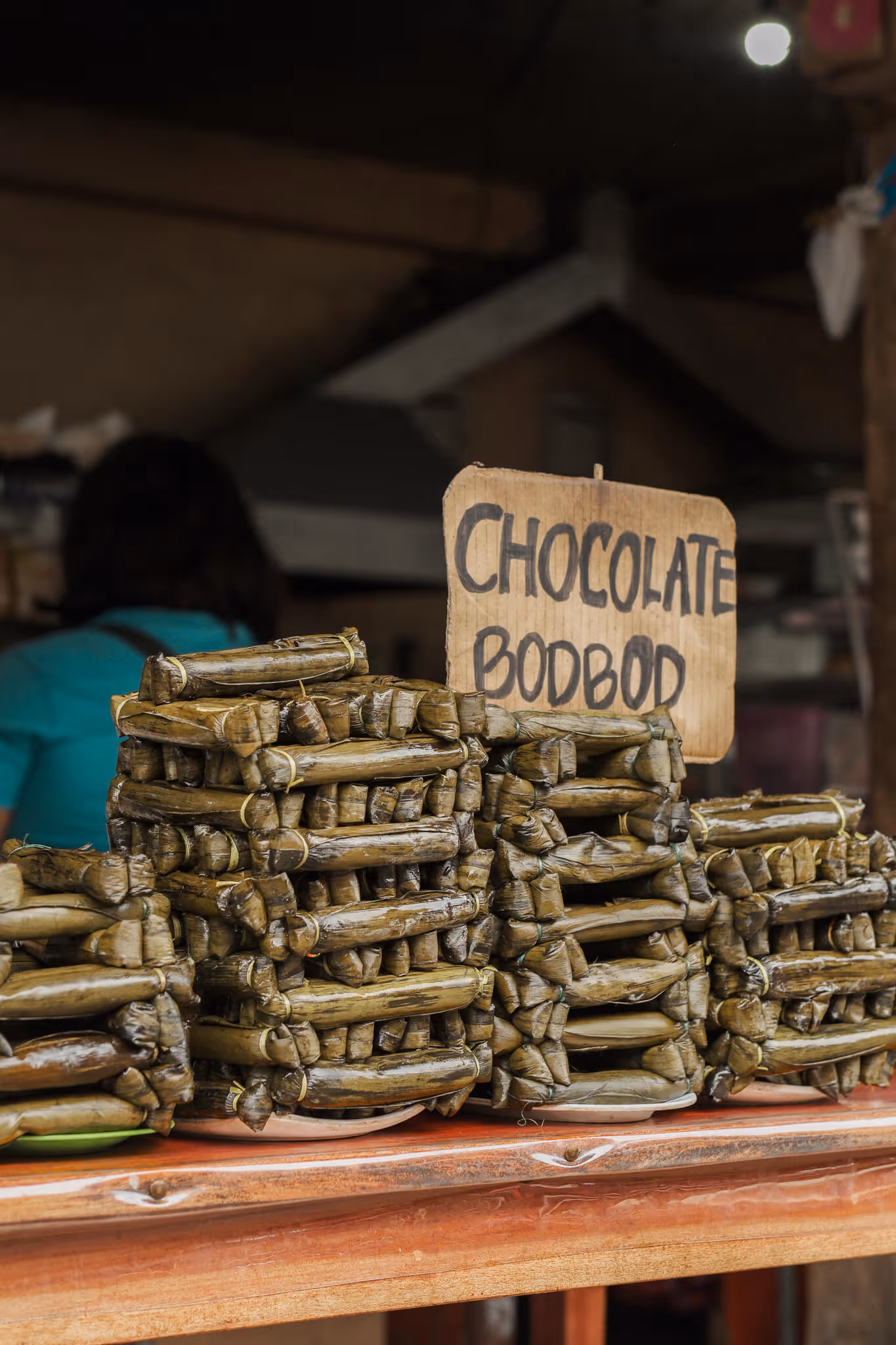 Stacks of chocolate bodbod wrapped in banana leaves on a wooden table in a market.