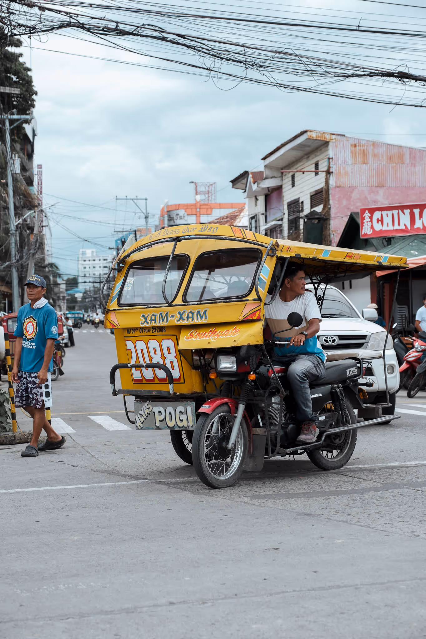 Man driving yellow tricycle taxi on a street with another man walking nearby.
