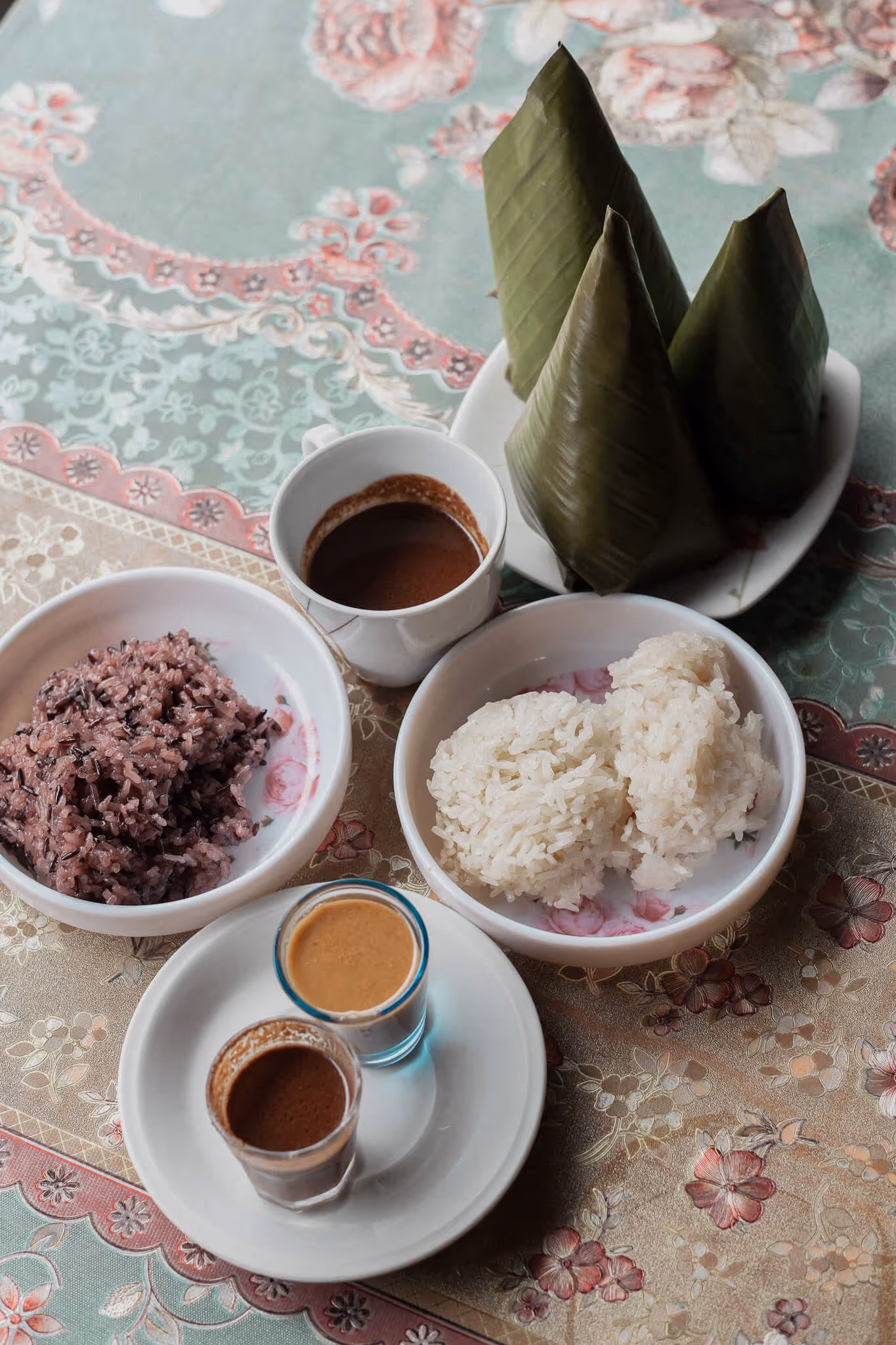 Bowls of sticky rice, cup of brown sauce, two small glasses of sauces on floral tablecloth.