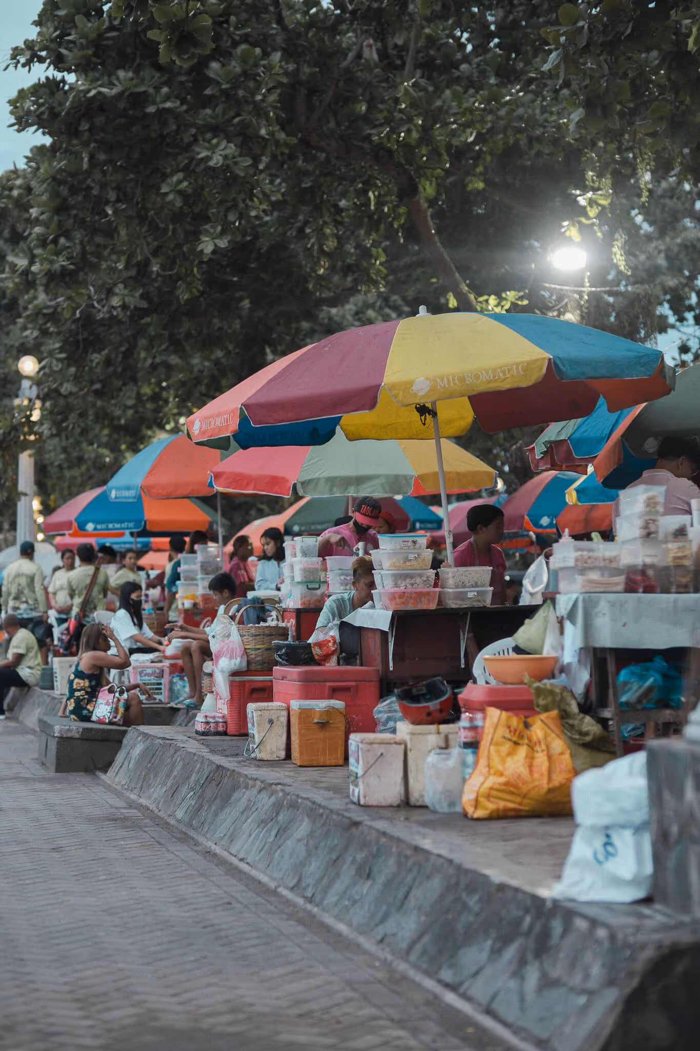 Street market stalls with colorful umbrellas and vendors selling goods along a tree-lined path.