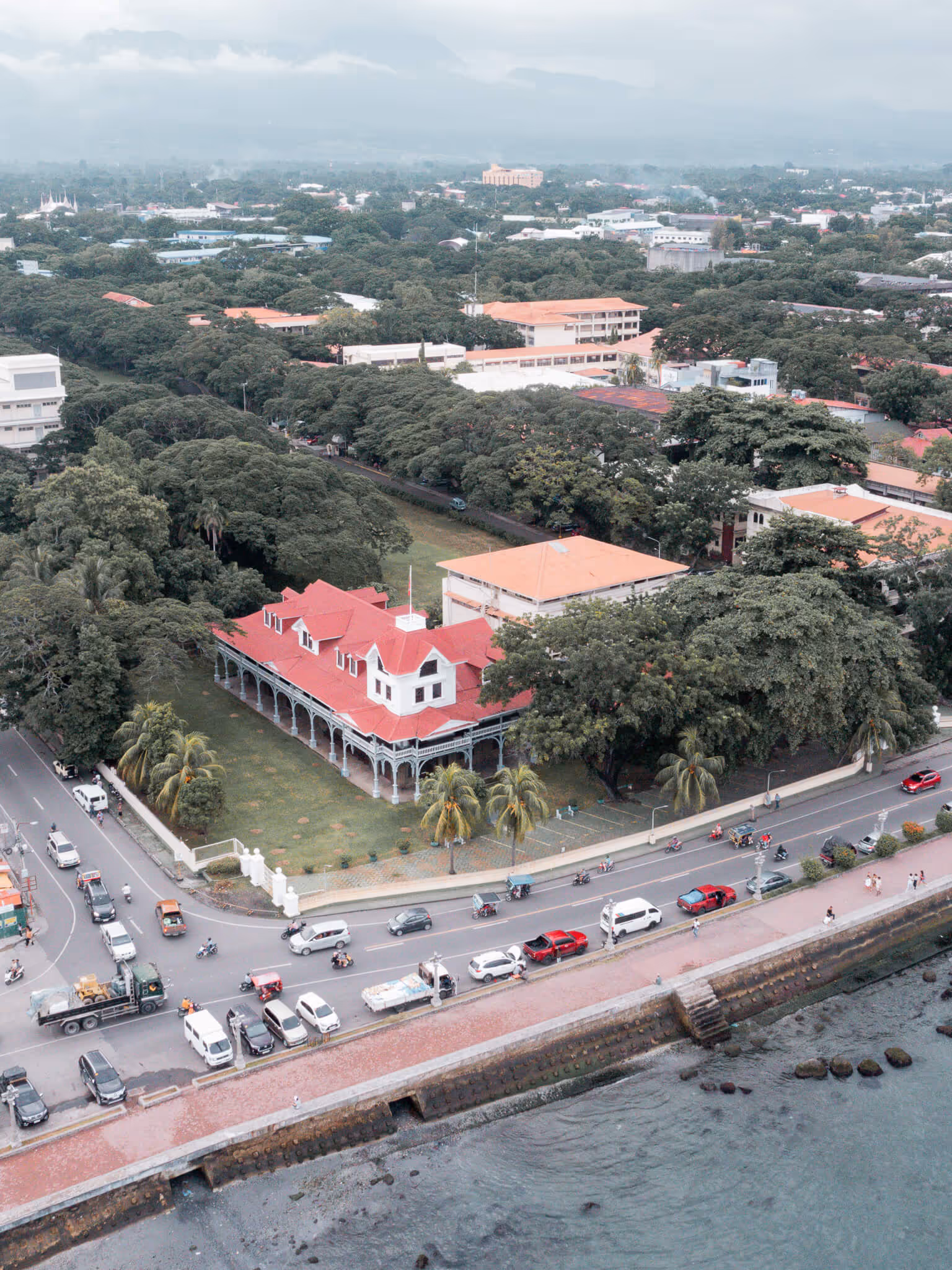 Aerial view of a red-roofed building beside a curved street with vehicles along a waterfront.