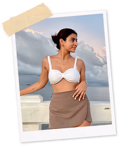 Angely Dub, Founder, in a white top and brown skirt stands by a waterfront under a cloudy sky.