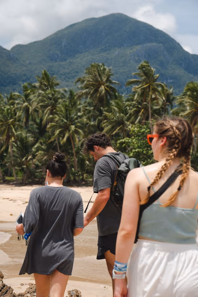 Three people walking on a beach with palm trees and a green mountain in the background.