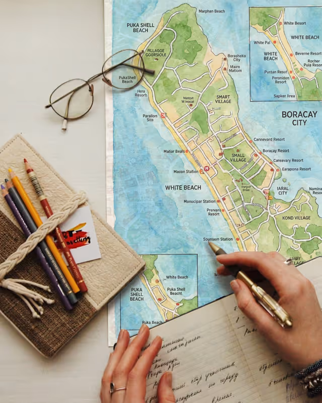 Person holding a pen over a Boracay map with notebooks, pencils, and glasses on a table.