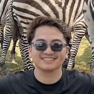 Smiling man wearing sunglasses and black shirt with zebras standing in background on grass.
