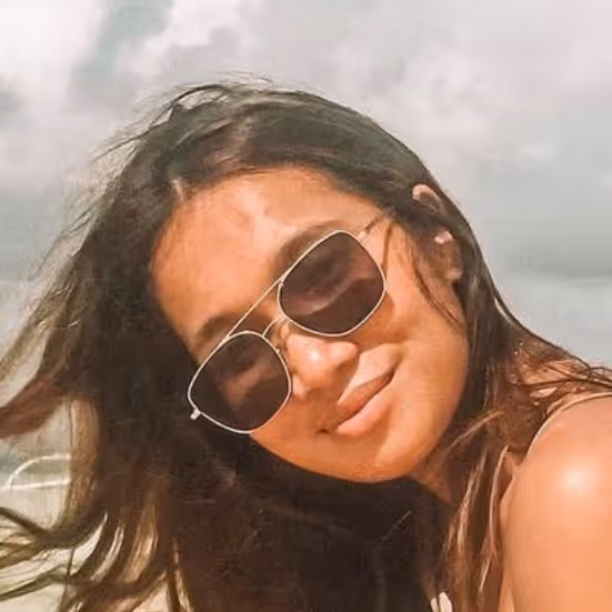Woman with long dark hair wearing sunglasses, smiling at the camera outdoors under a cloudy sky.