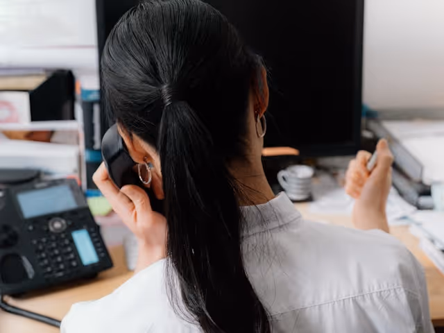 Woman with long black hair in a white shirt talking on a desk phone at her workspace.