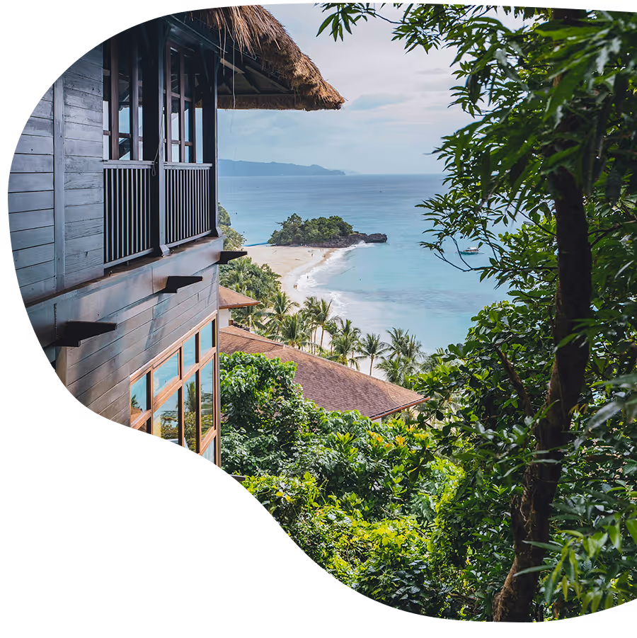 Wooden house beside lush trees overlooking a sandy beach and ocean with distant islands.