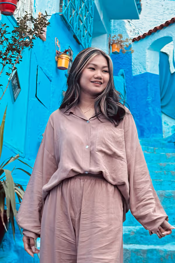 Smiling woman with dark hair wearing beige outfit standing in front of blue painted buildings and stairs.