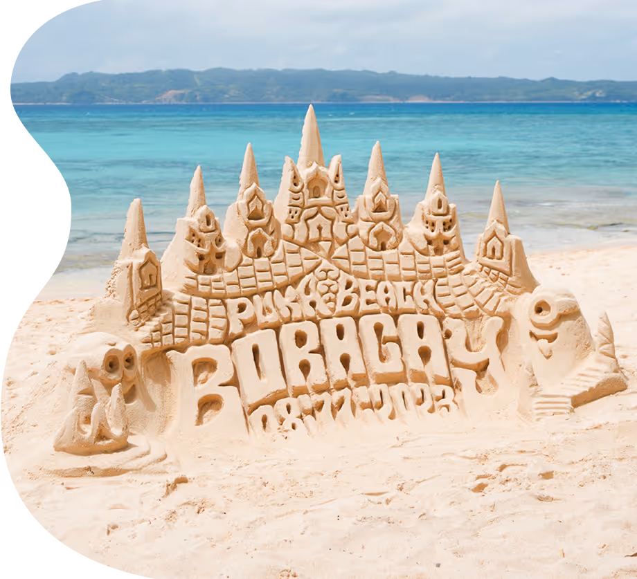 Intricate sandcastle on white beach with Boracay and Puka Beach carved, turquoise sea and hills behind.