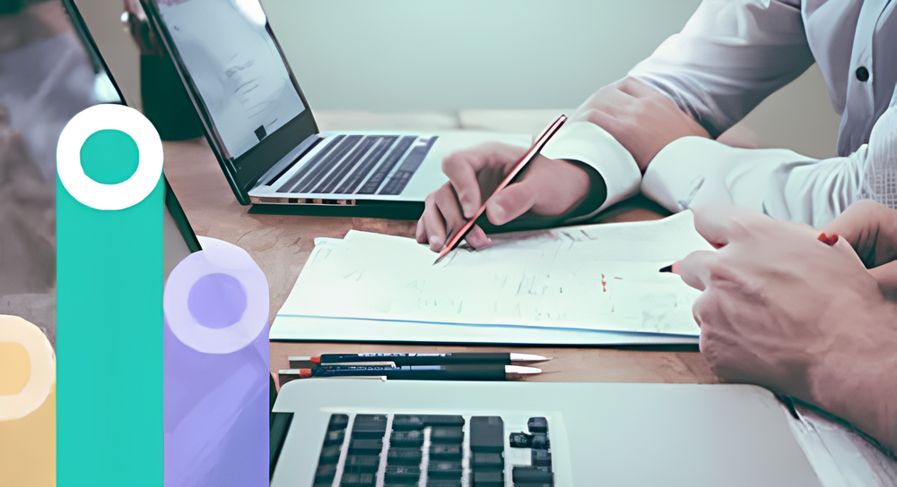 Two people working at a wooden table with laptops and papers, one writing with a pencil while the other points at the paper.