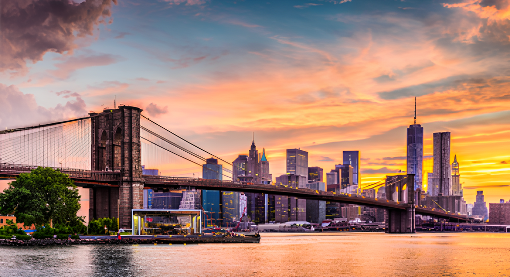 Brooklyn Bridge stretching across the East River with the Manhattan skyline and a colorful sunset sky in the background.