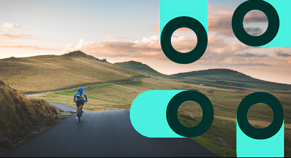 Cyclist riding on a paved road through gently rolling grassy hills under a partly cloudy sky at sunset, with abstract teal and dark green circular shapes overlaid on the right side of the image.