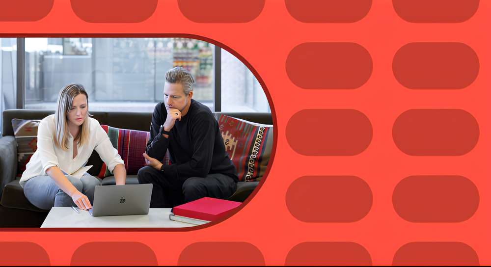 A man and a woman sitting on a couch looking intently at a laptop together on a white table in front of them, with colorful cushions behind them and a red book on the table.