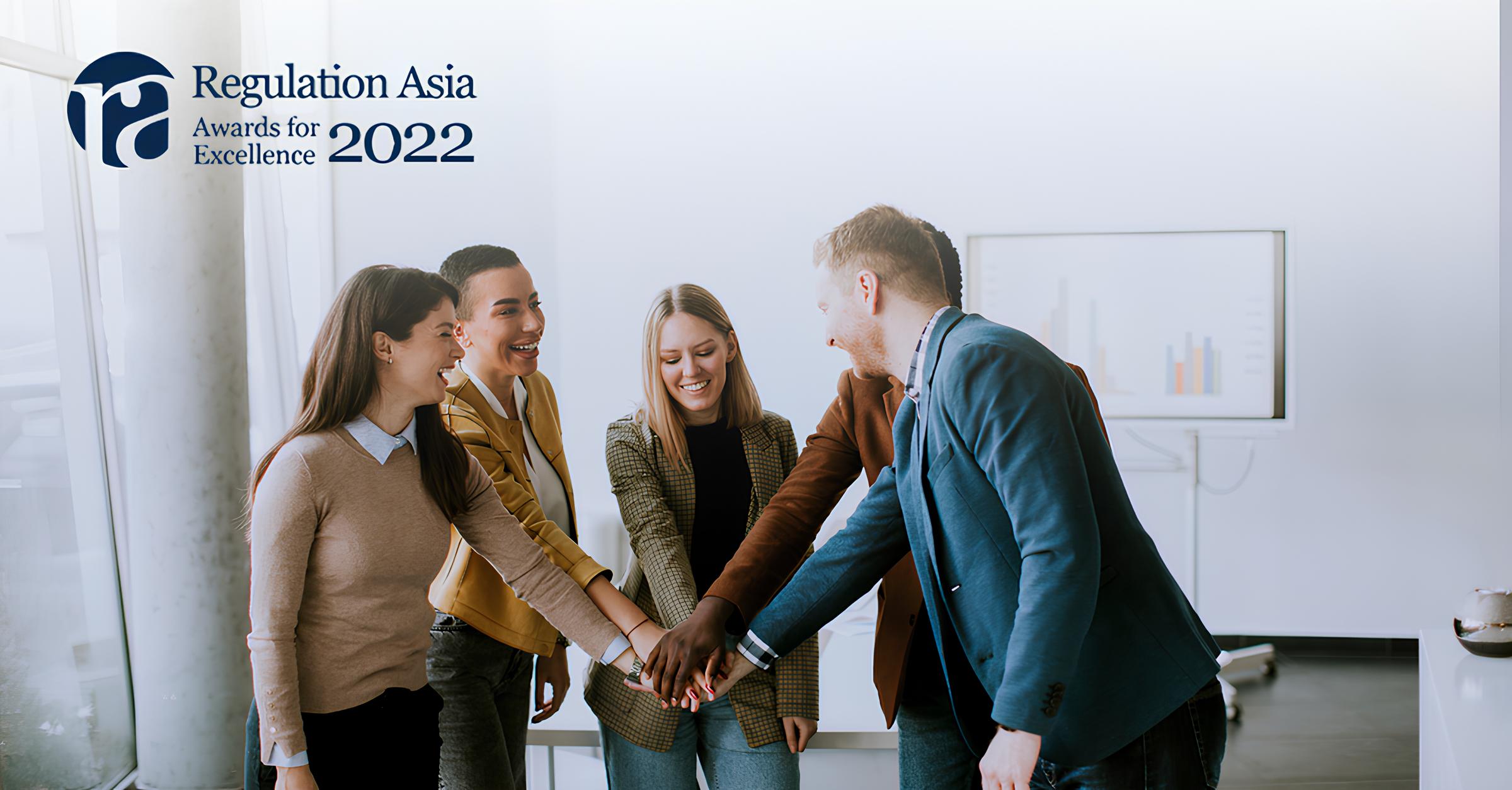 Five diverse professionals standing in a modern office with light walls and a window, joining their hands together in a teamwork gesture, smiling and happy, with a chart on a screen in the background and Regulation Asia Awards for Excellence 2022 logo in the top left corner.