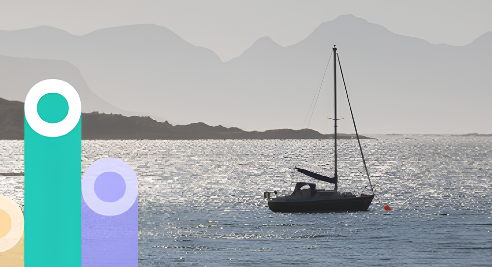 Sailboat anchored on calm water with sun reflecting on the surface and misty mountains in the background.