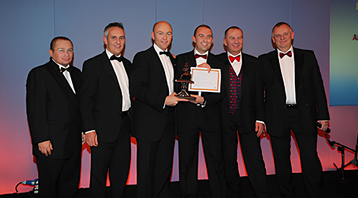 Six men dressed in black tuxedos with bow ties standing side by side on a stage, two of them holding an award and certificate, with a gradient blue and purple background.