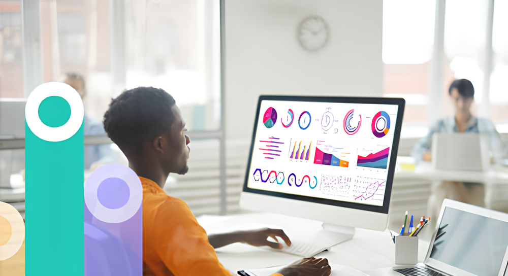Man in an orange shirt working at a desk with a large computer monitor displaying colorful charts and graphs, in an office with large windows and blurred background figures.