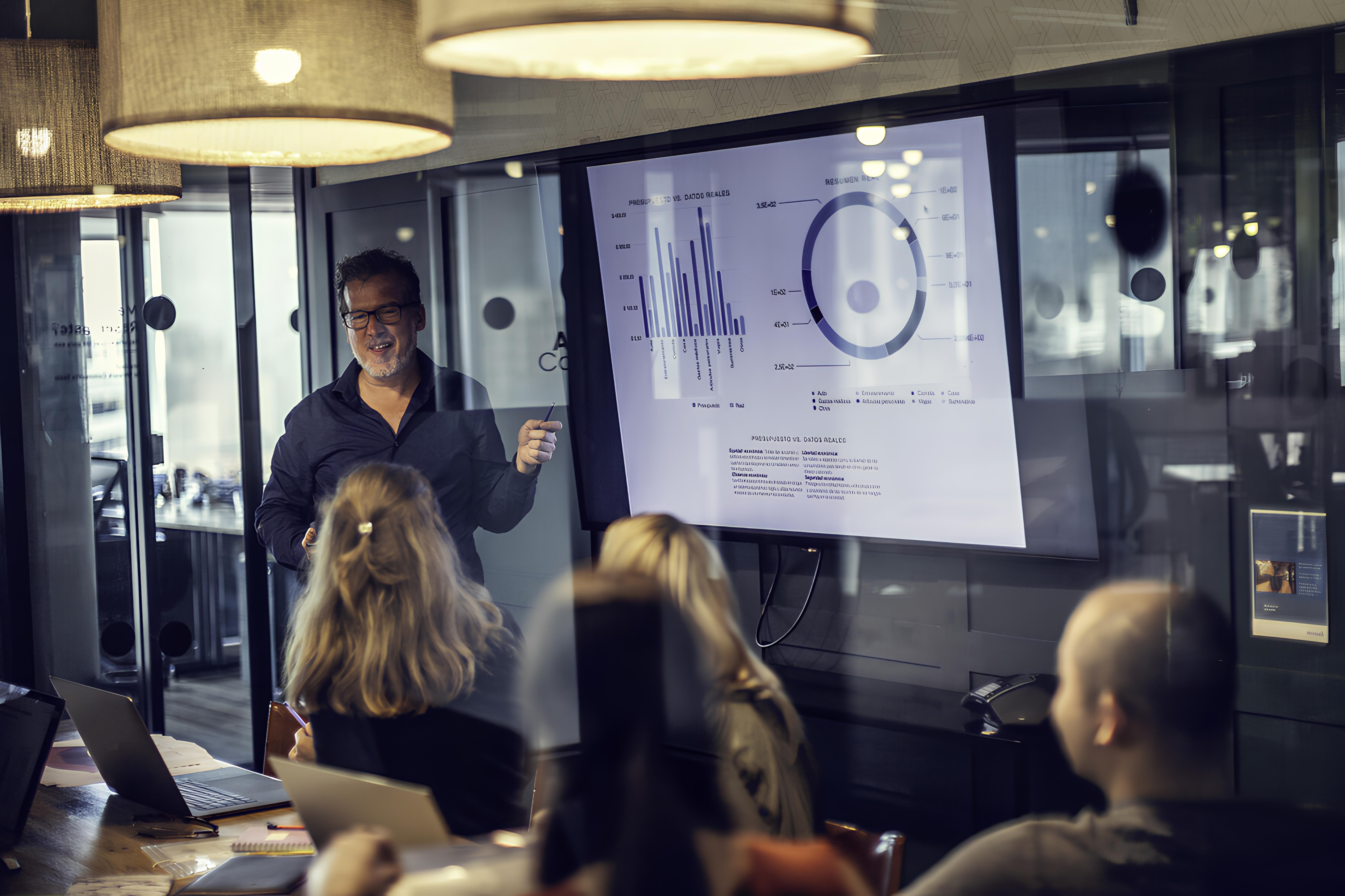 Man with glasses giving a presentation to a group in a modern office conference room, pointing at a large screen showing bar and pie charts with data.