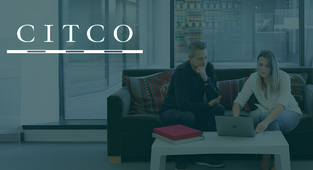 Two people sitting on a sofa in a modern office, focused on a laptop on a white coffee table with stacked books, with a large CITCO logo on the left side.