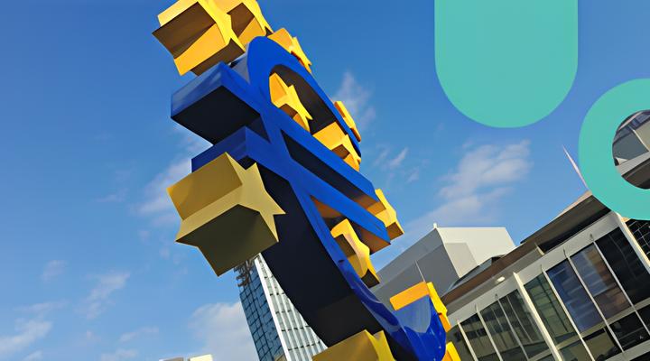 Large sculpture of a blue Euro currency symbol surrounded by yellow stars, set outdoors against a background of modern buildings and a blue sky with some clouds.