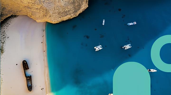 Aerial view of a shipwreck on a sandy beach with clear turquoise water and several small boats anchored nearby, bordered by a rocky cliff.