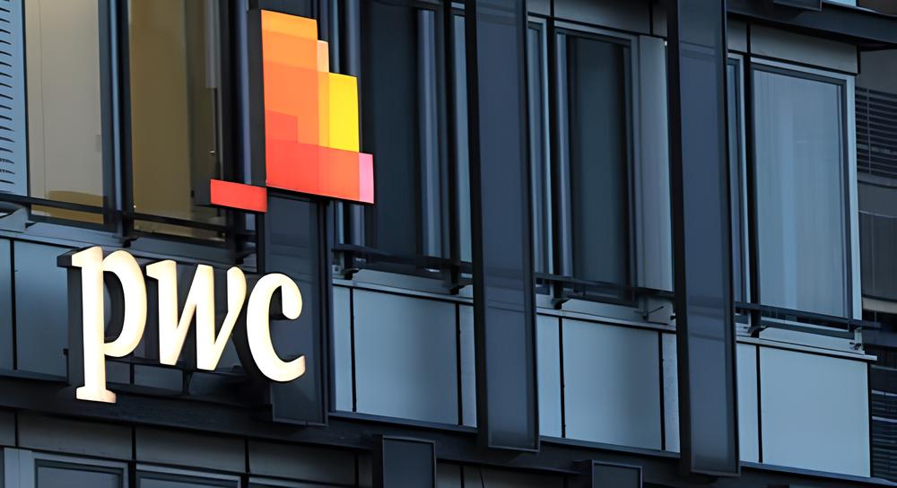 Illuminated PwC logo with red, orange, and yellow squares on a modern glass building facade at dusk.