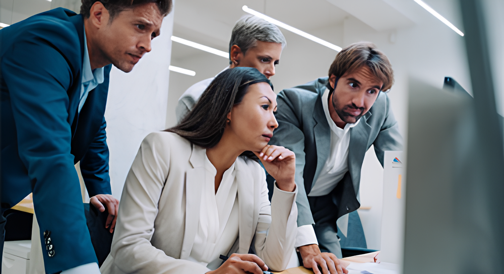 Four focused business professionals, two men and two women of diverse backgrounds, gathered closely around a computer screen in a modern office, engaged in a serious discussion or project.