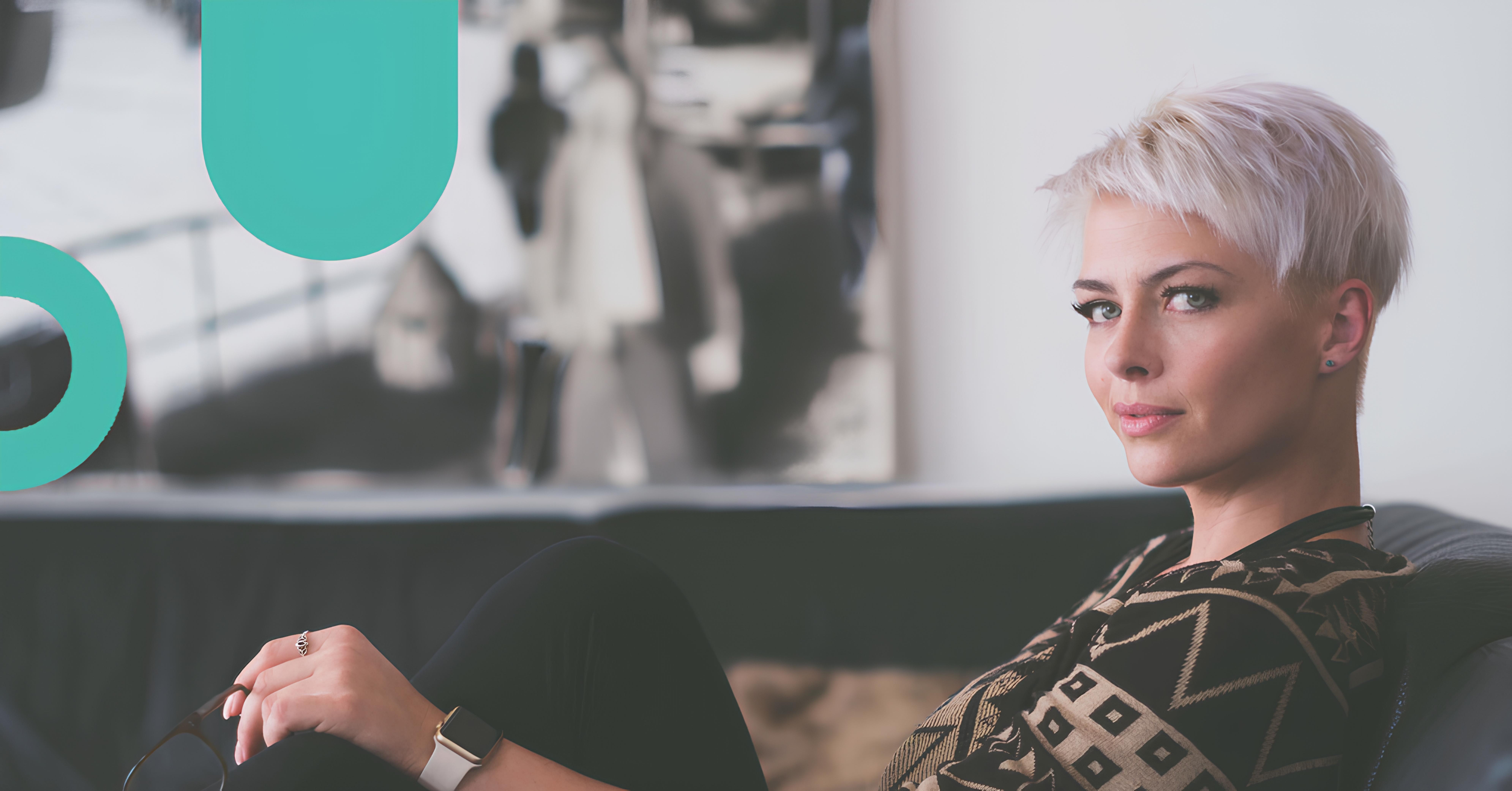 A woman with short platinum blonde hair and green eyes sits on a couch, looking towards the camera, wearing a black and gold patterned top, holding eyeglasses in one hand and wearing a smartwatch on her wrist.