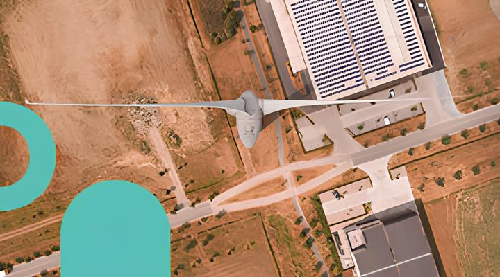 Aerial view of a wind turbine with three blades near buildings and roads, surrounded by dry, brown vegetation and some scattered small trees.