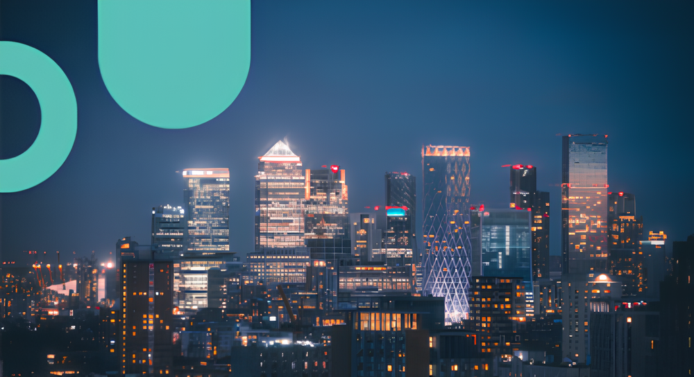 Nighttime city skyline with illuminated modern skyscrapers and high-rise buildings under a clear dark blue sky, featuring distinctive architectural lighting and some abstract teal graphic shapes in the corner.
