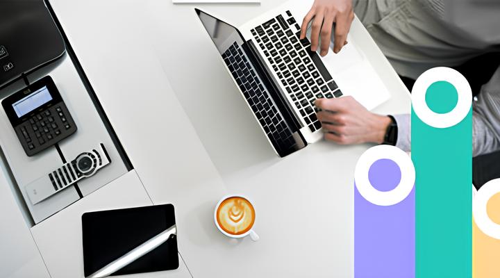 Top-down view of a person typing on a laptop at a white desk with a cup of latte coffee, a tablet with stylus, and office phone nearby.