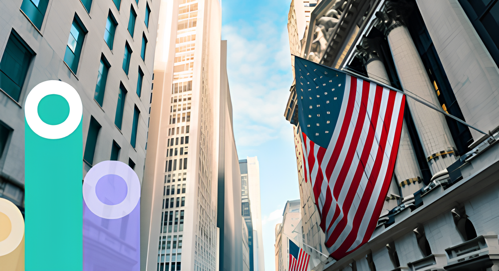 Street view of tall buildings with American flags hanging from the right side, under a partly cloudy blue sky.