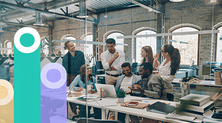 A group of seven diverse coworkers collaborating in a modern office with exposed brick walls and large arched windows, gathered around a desk with laptops, notebooks, and documents.