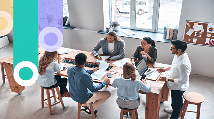 Six diverse colleagues gathered around a wooden table in a bright office, discussing documents and laptops during a meeting.