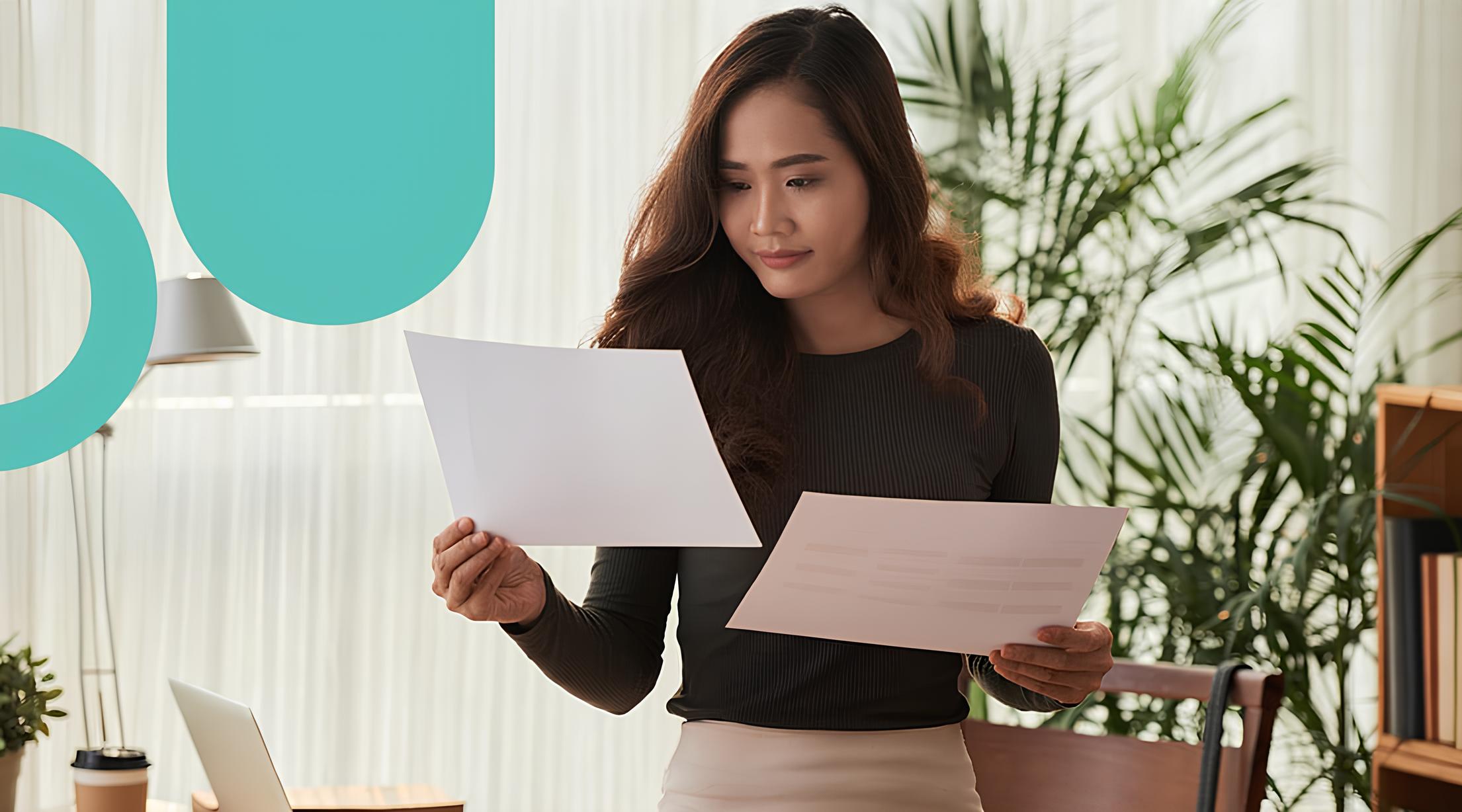 A woman with long brown hair wearing a black top and beige skirt stands indoors, reading two sheets of paper, with green plants and a laptop in the background.
