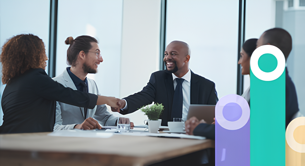 Diverse group of business professionals sitting around a conference table in a modern office, two of them smiling and bumping fists in greeting or celebration.