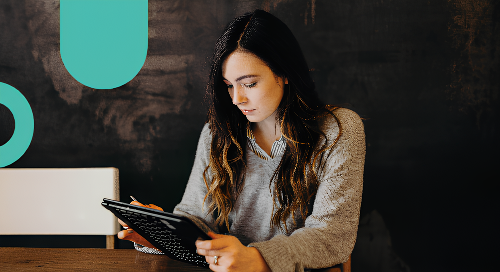 Young woman with long dark hair wearing a gray sweater is sitting at a wooden table and writing on a tablet with a pen, against a dark wall with teal abstract shapes.