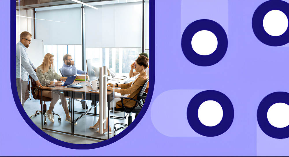 A group of five colleagues collaborating in a modern office conference room with glass walls, laptops, and desktop monitors, engaged in discussion and work.