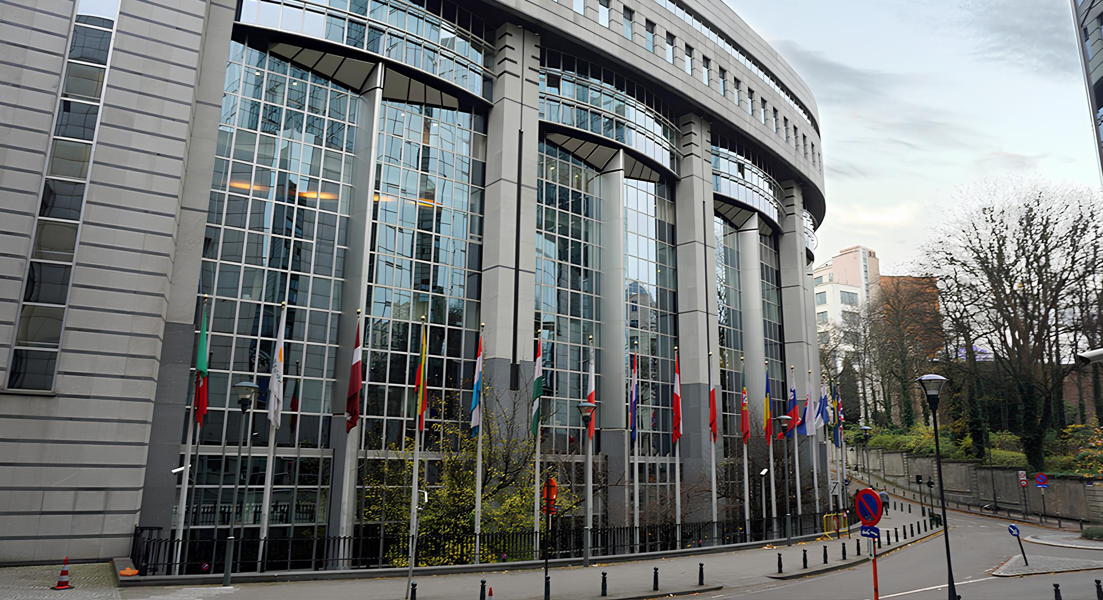 Curved modern government or institutional building with tall glass windows, a row of international flags on flagpoles in front, and a street with lampposts and traffic signs in an urban area with trees and other buildings nearby.