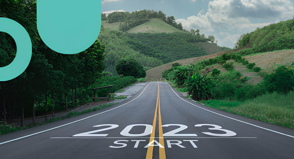 A paved road with double yellow lines running straight ahead through green hills and trees under a cloudy blue sky, featuring the year '2023' and the word 'START' painted on the road surface at the foreground.