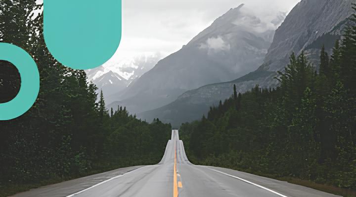 Empty two-lane road stretching straight ahead through dense green forest with large mountains and misty clouds in the background under a cloudy sky.