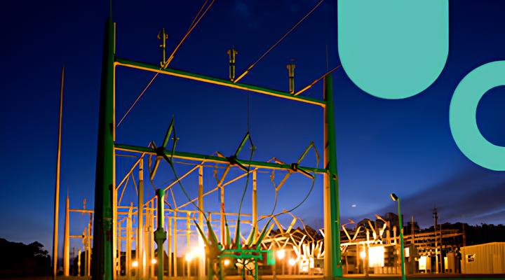 Electric power substation at dusk with green metal framework, cables, and bright lights against a deep blue sky.