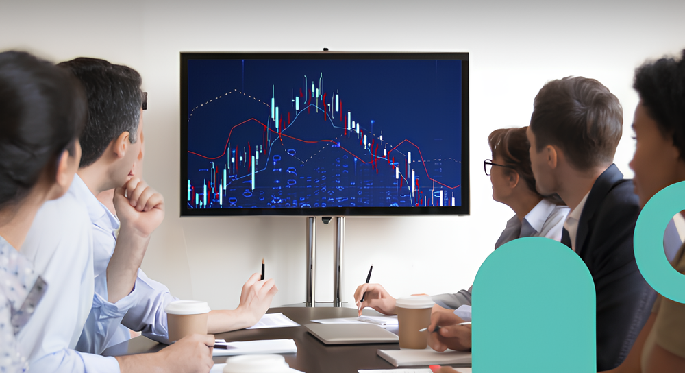 A group of five business professionals sitting at a table looking at a large screen displaying a financial candlestick chart with red and blue trend lines.