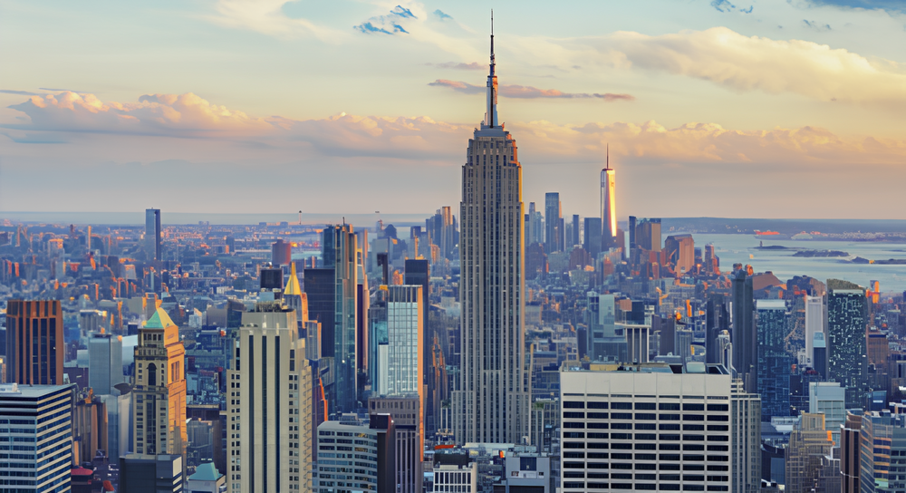 Panoramic view of New York City skyline at sunset featuring the Empire State Building centrally, surrounded by numerous skyscrapers with a partly cloudy sky above and water in the distant background.